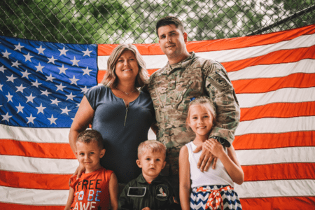 Military soldier next to his wife standing behind their three young children, with a large American Flag in the background .